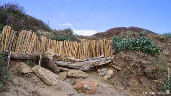 A rustic wooden barrier made of bamboo and driftwood encloses a sandy path leading to a coastal area, bordered by green vegetation and rocky elements under a clear blue sky.