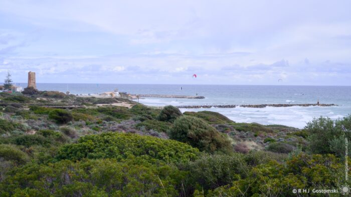 A scenic coastal view featuring a rocky shoreline, waves, and a cloudy sky. There are kite surfers in the distance, lush greenery in the foreground, and a historic tower on the left near the ocean.