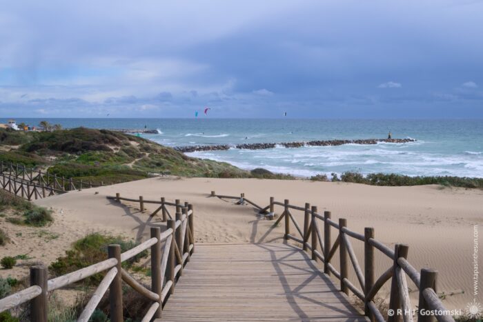 A wooden boardwalk blocked by sand dunes leads towards a beach with gentle waves and kitesurfers in the distance under a dramatic cloudy sky.
