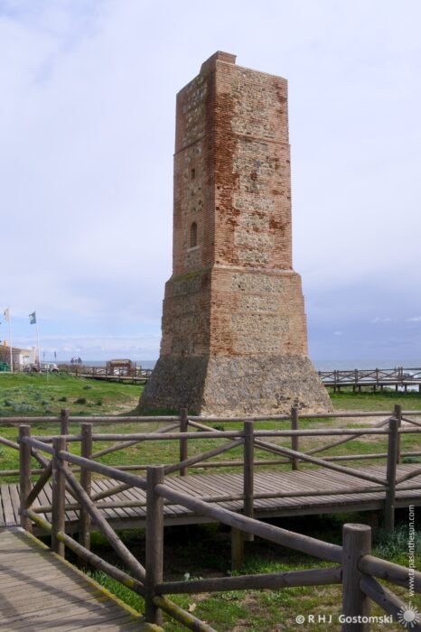 Torre Ladrones stands beside a wooden walkway, surrounded by greenery and near a body of water under a cloudy sky.