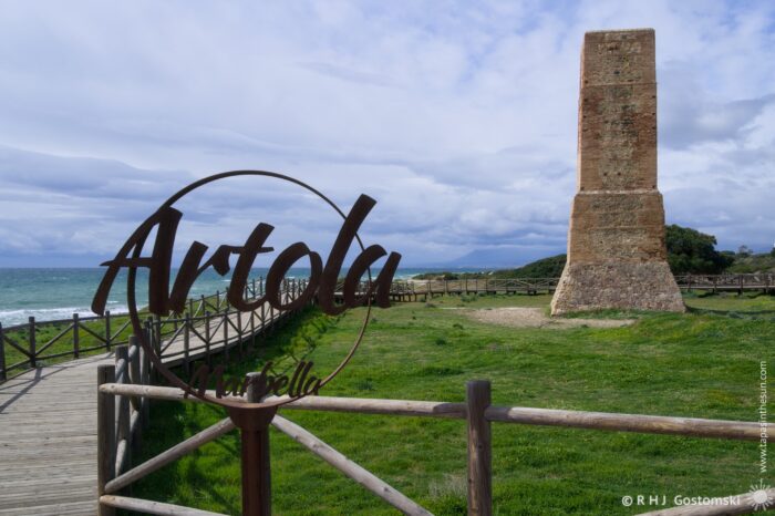 A coastal view featuring the Artola sign and the ancient Torre Ladrones watchtower, surrounded by green grass and a wooden walkway leading towards the sea under a partly cloudy sky.