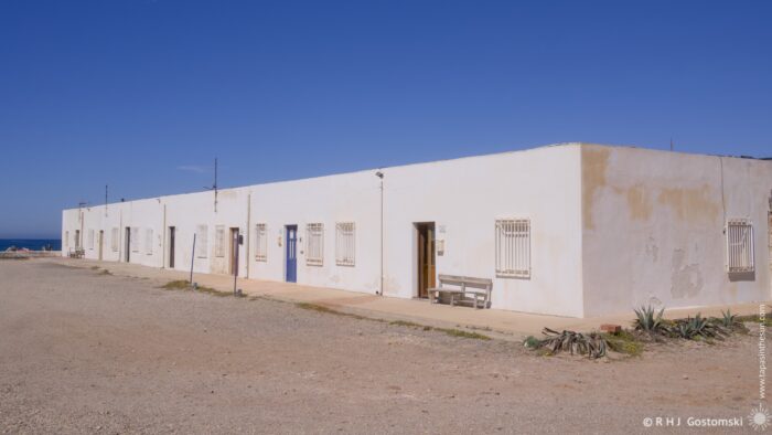 A row of whitewashed houses in Las Salinas Workers&rsquo; Village stretch along a paved walkway and gravelly road. A calm blue sea is visible in the distance. The architecture is simple and utilitarian, with small windows and doorways creating a sense of quiet solitude. A few plants add touches of green to the otherwise monochromatic scene.