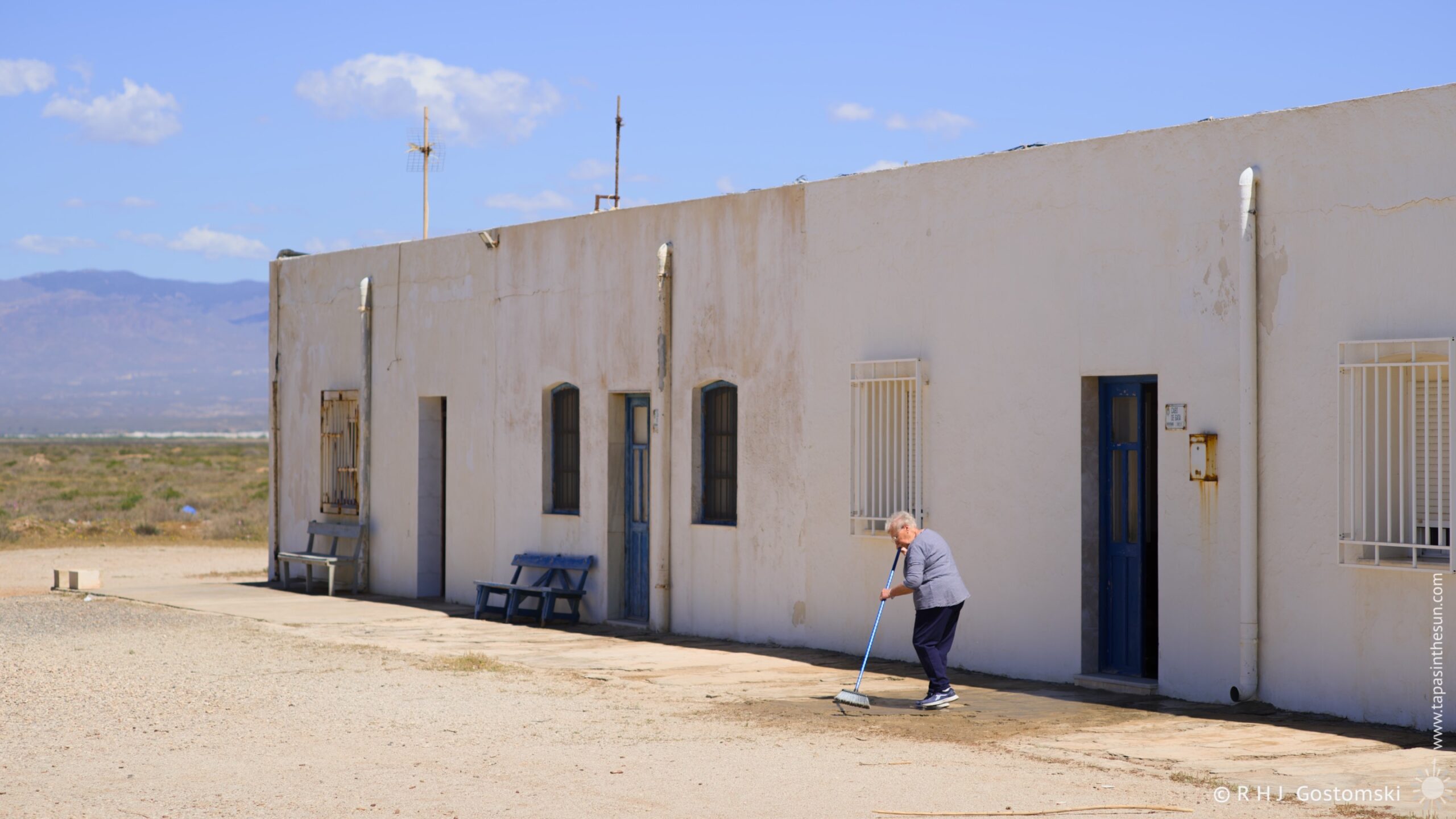 A woman meticulously sweeps a gravel courtyard in front of a row of whitewashed houses in Las Salinas Workers’ Village under a bright, midday sun. The scene is set against a backdrop of an arid landscape and distant mountains, creating a sense of quiet solitude. The composition emphasises the woman's activity as he tends to her surroundings.