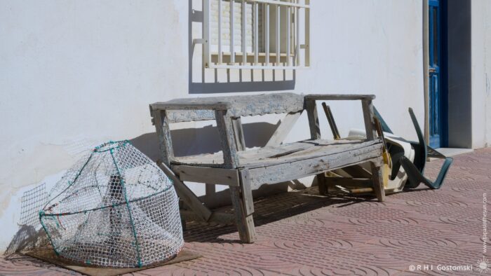 A weathered wooden bench sits on a tiled patio outside a whitewashed building in Las Salinas, Cabo de Gata. A fishing net and overturned chairs are scattered nearby. The bright midday light casts strong shadows across the scene.