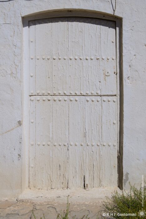 A stable style door with a curved top is set into a textured white stone wall. The wooden door, adorned with rivets, appears weathered and aged, creating a sense of history and simplicity. The midday light highlights the texture of both the wall and the shutter.