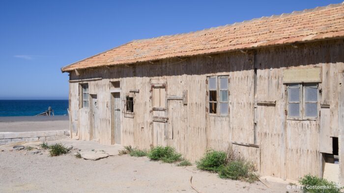 A long, weathered wooden warehouse building with multiple windows and a tiled roof sits beside a gravelly yard and sandy beach. The structure appears abandoned, showing signs of age and exposure to the elements, with the sea visible in the background. The bright sunlight suggests a mid-morning scene.