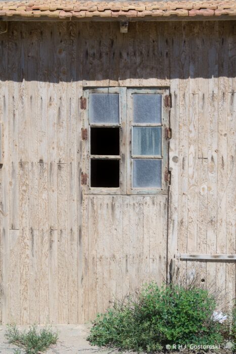 A close-up view of an old, weathered wooden wall in Las Salinas, Cabo de Gata. A small window with six panes, two of which are missing, sits within the wall. The scene evokes a sense of quiet solitude and aged charm.