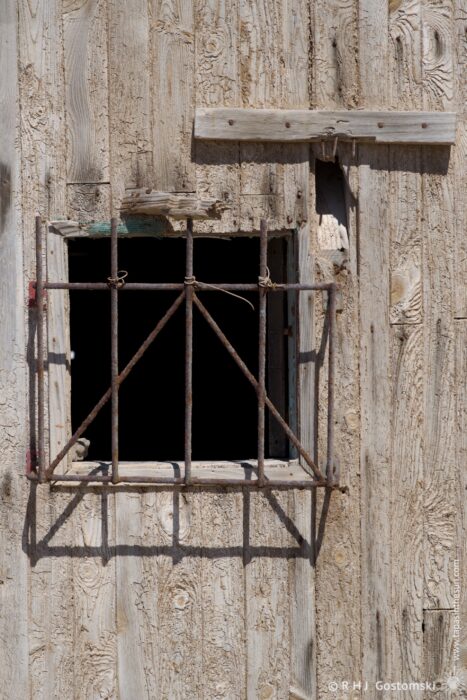 A weathered wooden building facade features a square window framed by rusty metal bars, creating an intriguing interplay of light and shadow. The rough texture of the wood contrasts with the geometric pattern of the window, evoking a sense of history and abandonment. The midday sun casts stark shadows across the surface.
