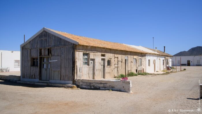A long, weathered wooden building stands in stark contrast to the bright blue sky and arid landscape. The structure appears abandoned, with boarded-up windows and a faded roofline, evoking a sense of history and decay. The scene is bathed in the bright light of midday.