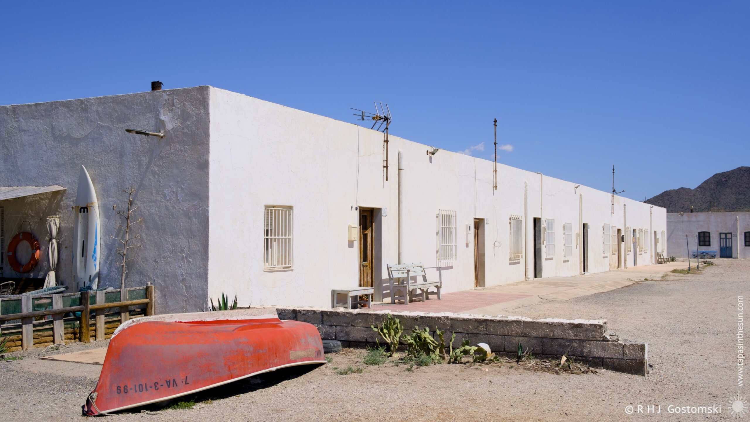 A long, whitewashed row of houses stretches along a gravel driveway in Las Salinas, Cabo de Gata. A small boat and surfboard propped against the building's exterior, adding a touch of coastal charm. The bright afternoon light illuminates the scene, creating a warm and inviting atmosphere.