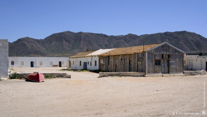 A weathered, low-lying building complex sits on a dusty ground in front of a range of arid mountains under a clear, bright sky. A small boat rests near the buildings, and the scene evokes a sense of solitude and abandonment.