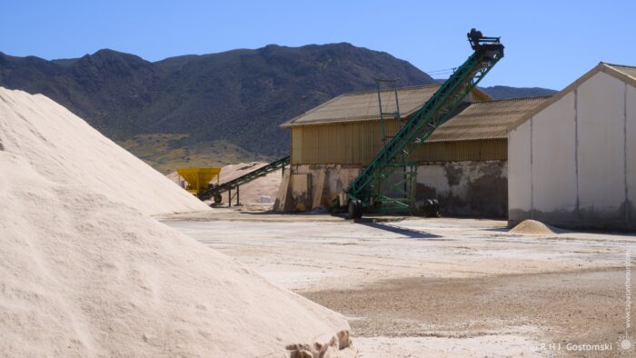 A large conveyor belt used for transporting salt stands in the yard at Las Salinas Salt Works. A series of buildings and piles of salt dominate the scene against a backdrop of distant, arid mountains under bright midday sunlight. The composition emphasises the industrial scale of salt extraction against a natural landscape.