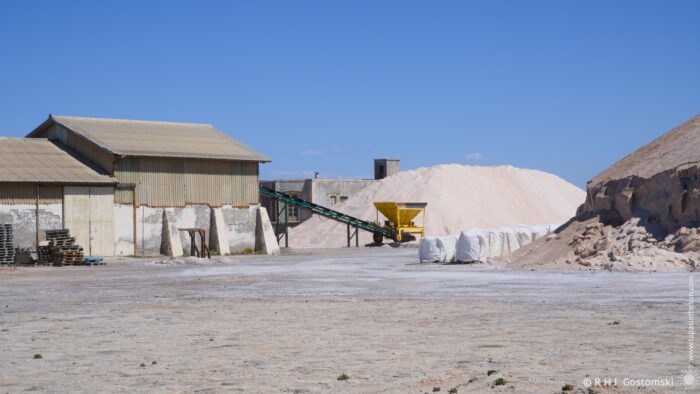A wide shot depicts an industrial salt harvesting operation on a bright, sunny day. Large piles of white salt dominate the foreground, with conveyor belts and processing buildings visible in the mid-ground against a clear blue sky. The scene conveys a sense of scale and industry.