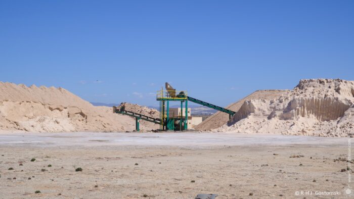 A large conveyor belt system dominates a barren landscape with huge stockpiles of salt, set against a clear blue sky. The scene evokes an industrial feel, with the machinery contrasting sharply with the natural environment. The bright sunlight creates strong shadows and highlights across the terrain.