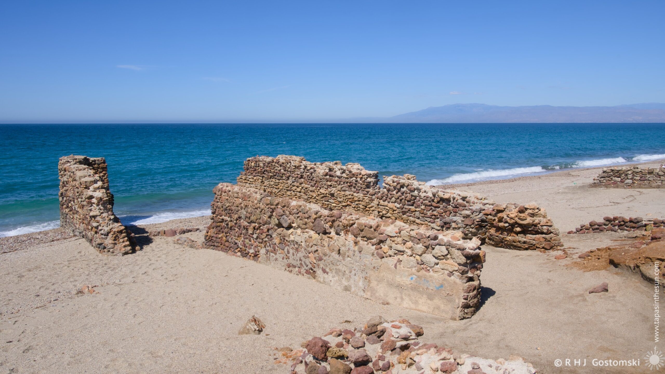 A weathered stone structure stands on a sandy beach, overlooking the turquoise waters of the Mediterranean Sea. Only remnants of walls remain of the salt-loading pier. A distant mountain range is visible across the sea on the horizon under a clear blue sky.