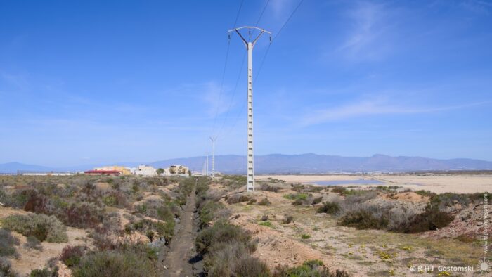 A water channel and row of power transmission pylons run into the distance, leading to the Las Salinas Salt Works and Workers&rsquo; Village in the distance. The midday sun blazes above, highlighting the arid terrain.