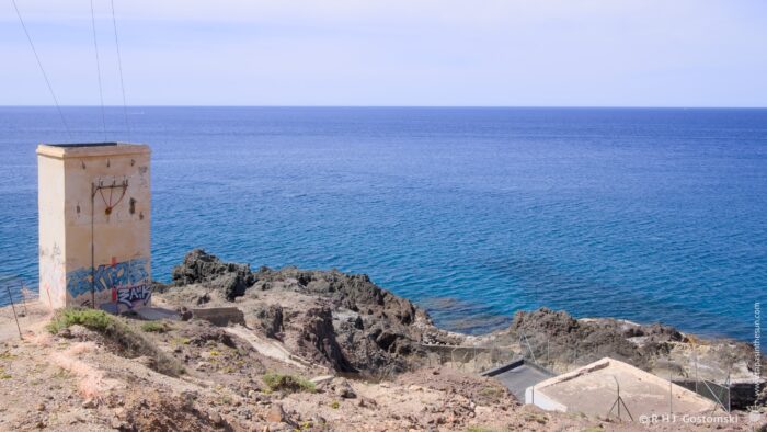 A weathered concrete sub-station stands on a rocky outcrop overlooking the deep blue sea, with dark volcanic rock formations below. The scene is bathed in bright midday sunlight, creating a stark contrast between the grey tower and the vibrant water. The composition emphasises the isolation of the structure against the vastness of the ocean.