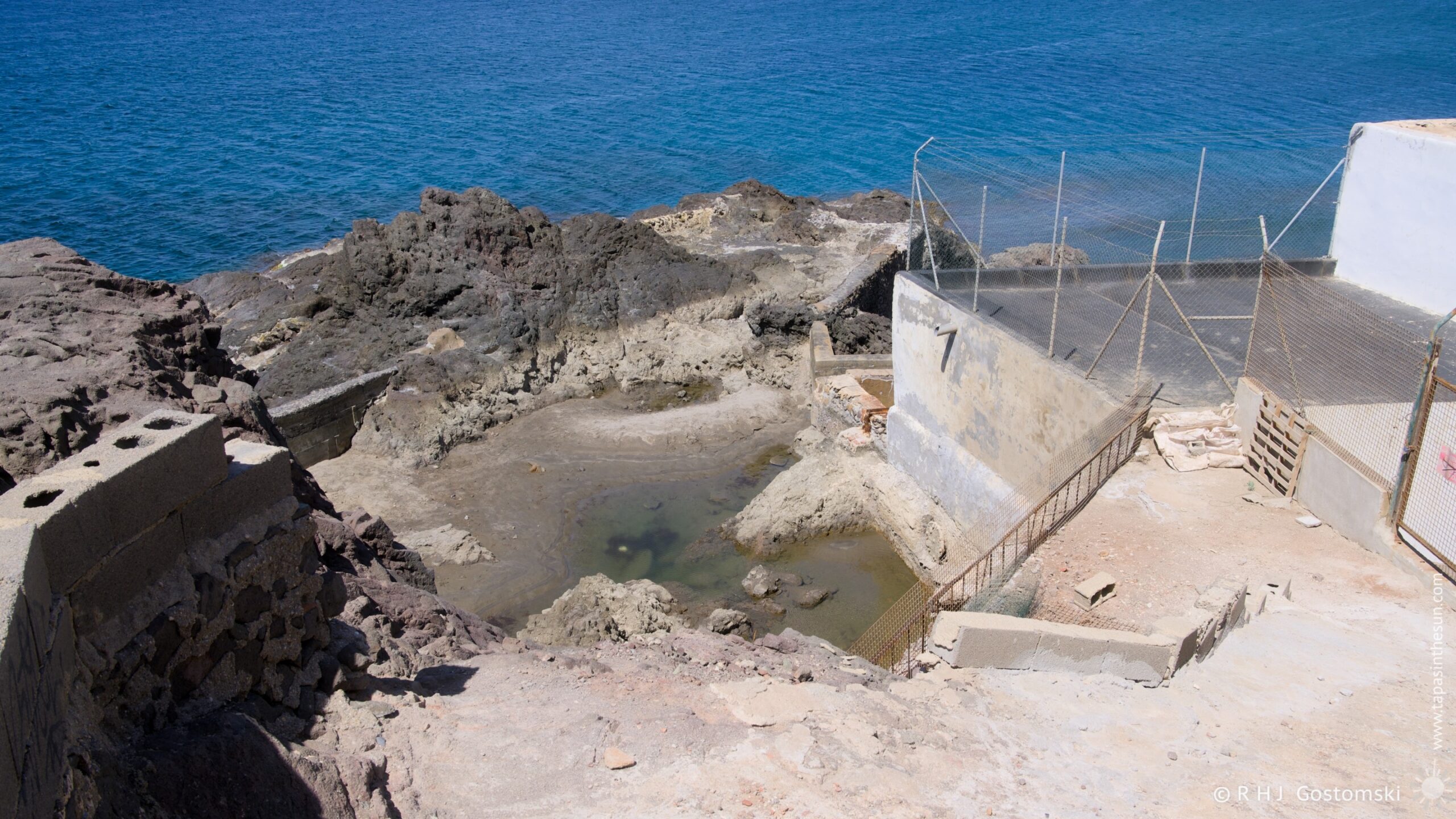 A high-angle view reveals a series of man-made pools nestled amongst dark, jagged rocks along the coast. Concrete structures and walkways frame the scene, with a weathered aesthetic enhanced by the bright midday light. The turquoise water of the pools contrasts sharply with the grey tones of the rock and concrete.