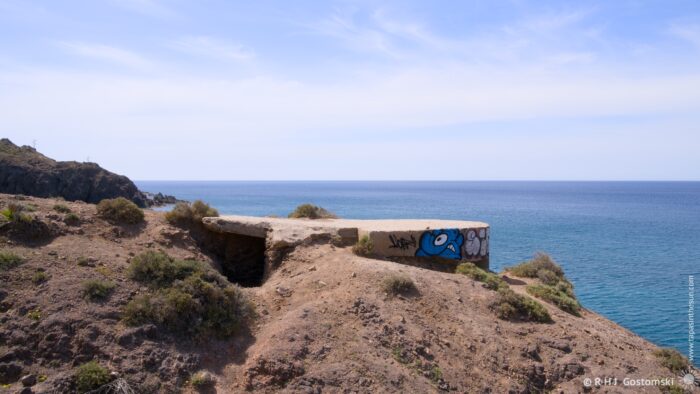 A concrete bunker, adorned with graffiti, sits perched on a rocky outcrop overlooking the turquoise waters of the Mediterranean Sea. The bright midday sun illuminates the scene, creating a clear and expansive view across the coastline. A sense of solitude and rugged beauty pervades the landscape.