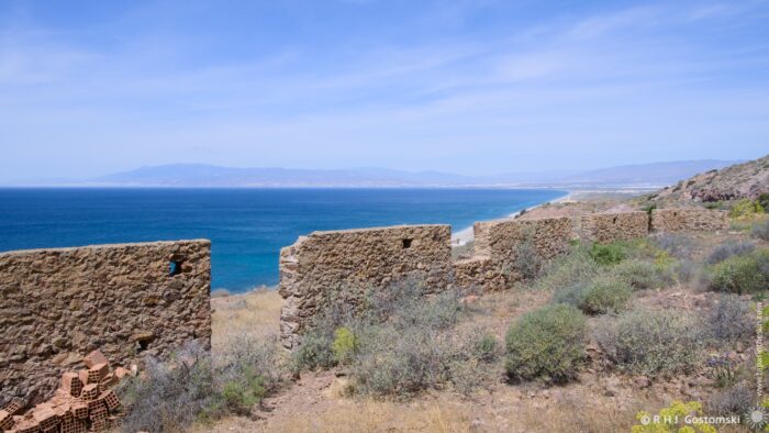 A weathered stone structure, partially collapsed, sits atop a hillside overlooking the deep blue sea and distant mountains under a clear sky. The foreground features dry, scrubby vegetation with touches of yellow flowers, creating a stark contrast against the ancient stonework and expansive landscape. The midday light casts strong shadows, highlighting the texture of the ruins.