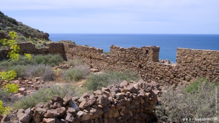Ruined stone walls, likely remnants of an old structure, cascade down a hillside towards the shimmering Mediterranean Sea. Sparse vegetation clings to the rocks and slopes, creating a textured foreground against the expansive blue horizon under a bright midday sun. The scene evokes a sense of history, resilience, and natural beauty.