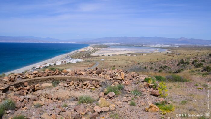 A wide, panoramic view from a rocky hillside overlooking the coastal landscape of Las Salinas de Cabo de Gata Salt Works. Along the coastline, a long stretch of sandy beach meets the turquoise sea, with salt flats and a small town visible in the distance, framed by rolling mountains under a bright afternoon sun.