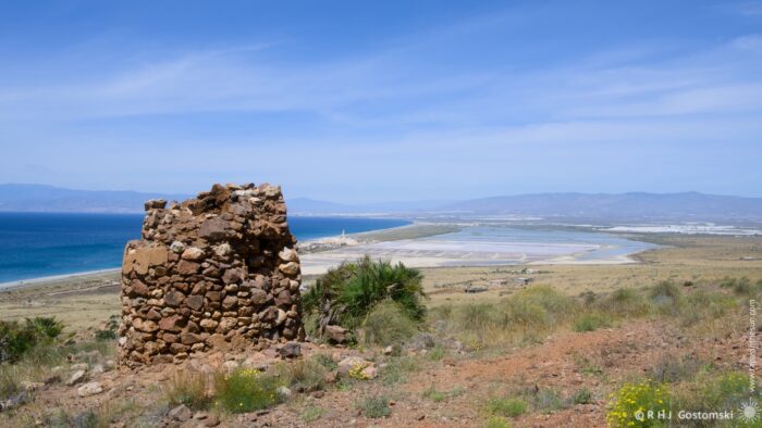 A weathered stone structure stands on a hillside, overlooking expansive salt flats and the distant sea under a bright afternoon sky. The scene evokes a sense of history, solitude, and natural beauty, with the composition drawing the eye from the foreground ruins to the expansive landscape.