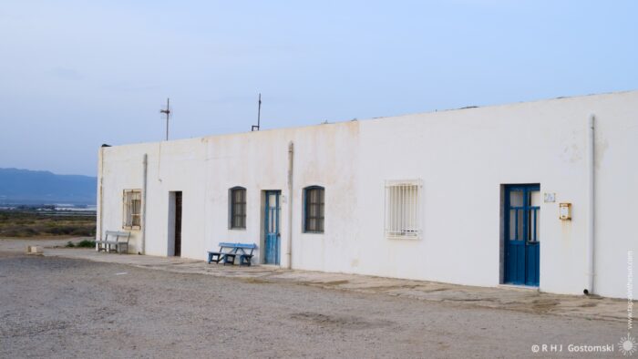 A long, white building with blue doors and windows faces a gravel expanse under the soft light of early evening. Two weathered benches sit in front, adding to the scene's tranquil atmosphere. Distant mountains are visible on the horizon.
