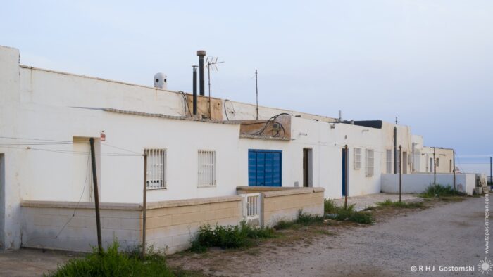A row of whitewashed houses with blue doors and windows lines a dusty road, facing the sea under a dusky evening sky. The scene evokes a sense of quiet solitude and timelessness, with the low light casting long shadows across the ground. The composition is dominated by horizontal lines and a muted colour palette.