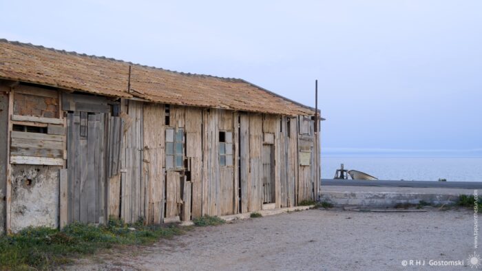 A weathered, abandoned warehouse with a tiled roof sits beside a grey beach under an overcast evening sky. A small wooden structure is visible in the distance, with the sea stretching out to the horizon. The scene evokes a sense of solitude and decay.