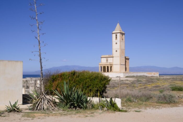 A tall, slender agave plant stands beside a low wall, with the Iglesia de las Salinas church in the distance across a flat landscape. The bright sunlight illuminates the scene on a clear day, creating a sense of tranquility and openness.