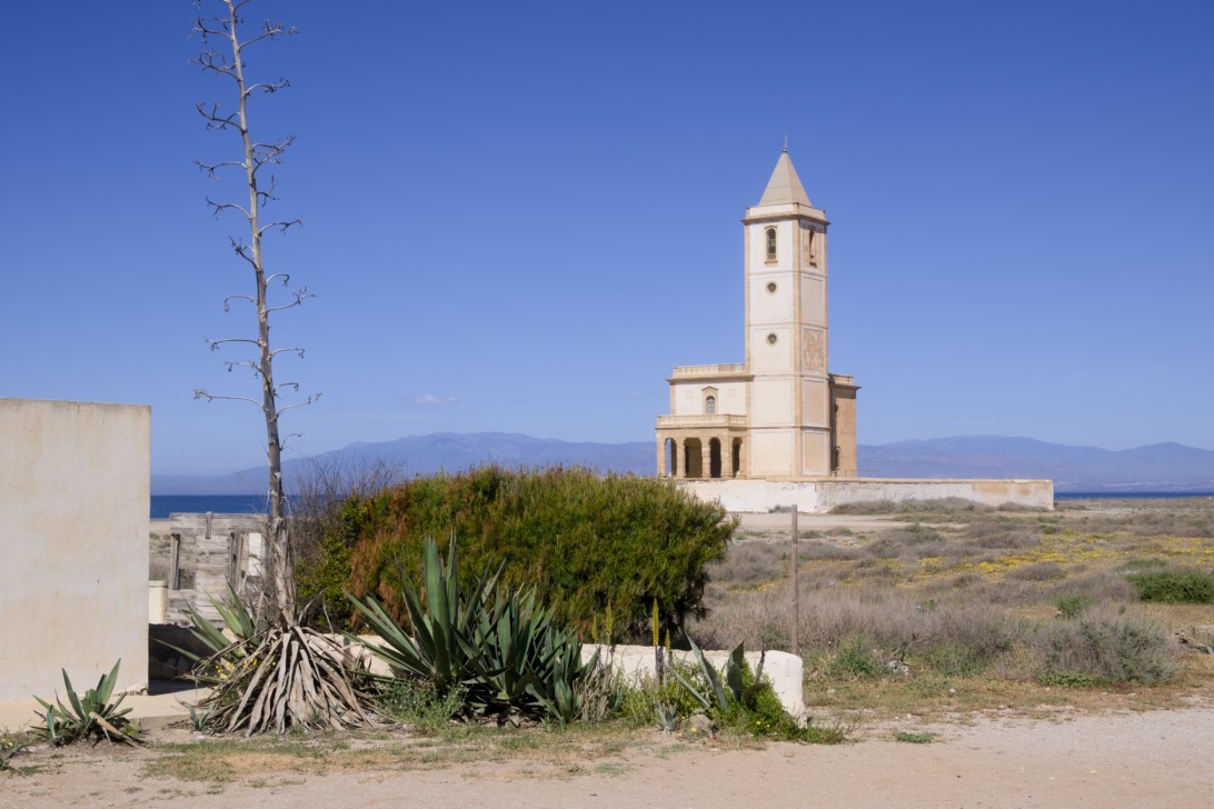 A tall, slender agave plant stands beside a low wall, with the Iglesia de las Salinas church in the distance across a flat landscape. The bright sunlight illuminates the scene on a clear day, creating a sense of tranquility and openness.